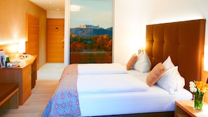 Quarto de hotel com uma cama grande, uma mesa com abajur e vista para um castelo através de uma porta de vidro.