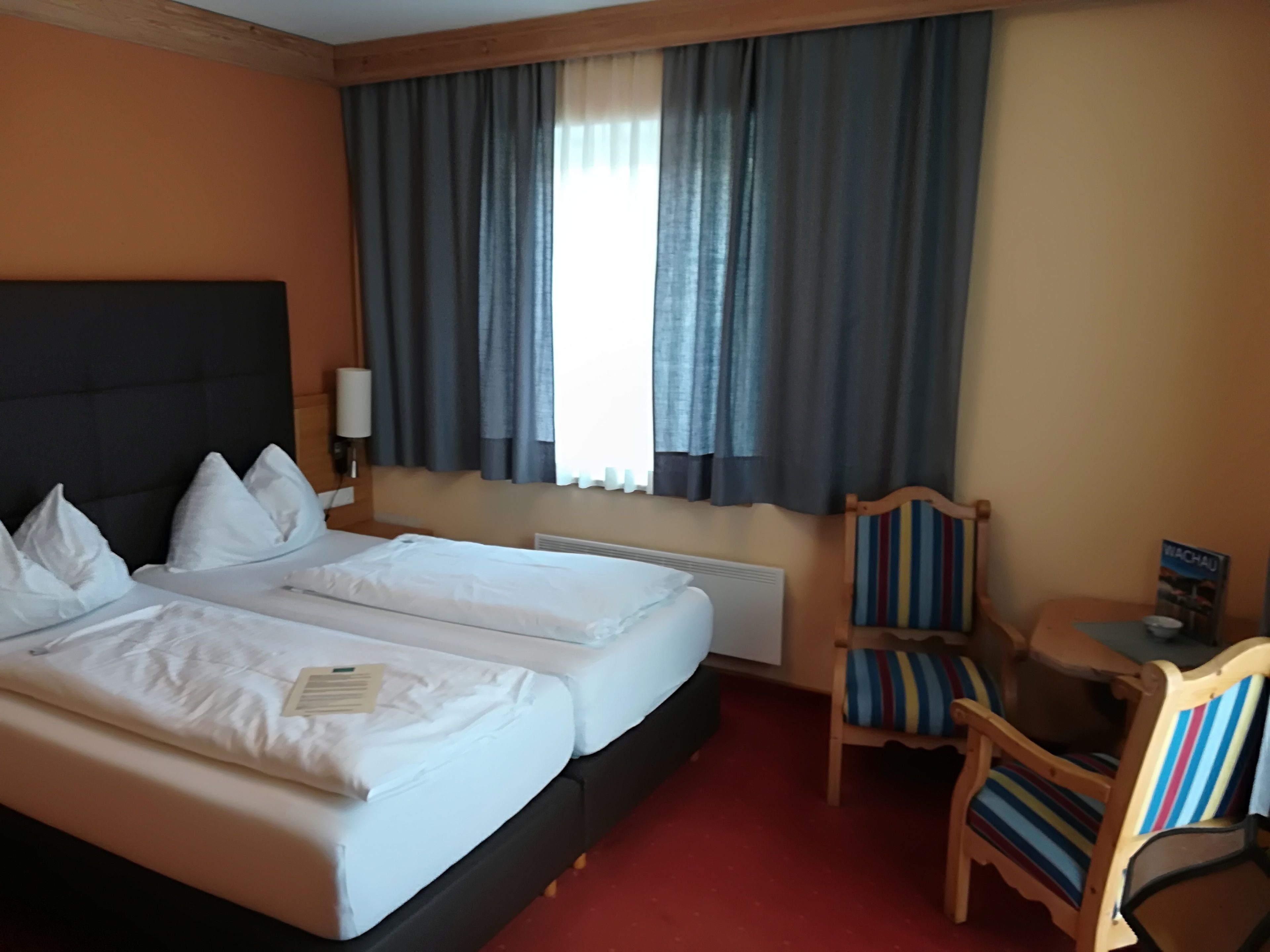 A hotel room with two beds, a wooden chair, a small table, and a window with curtains.