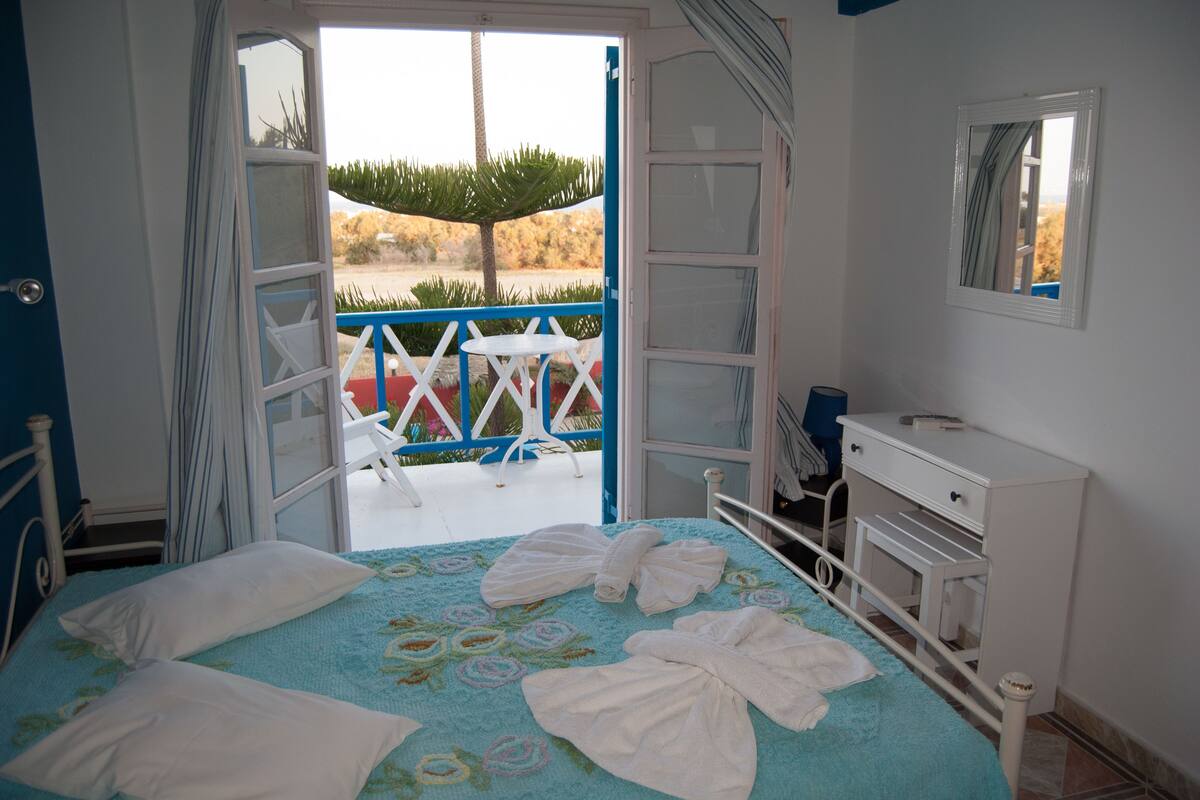 A bedroom with a bed, a dresser, and a balcony with a view of a palm tree and a fence.