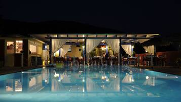 Ein Poolbereich bei Nacht mit überdachter Sitzgelegenheit und einem Gebäude mit Bar und Essbereich.