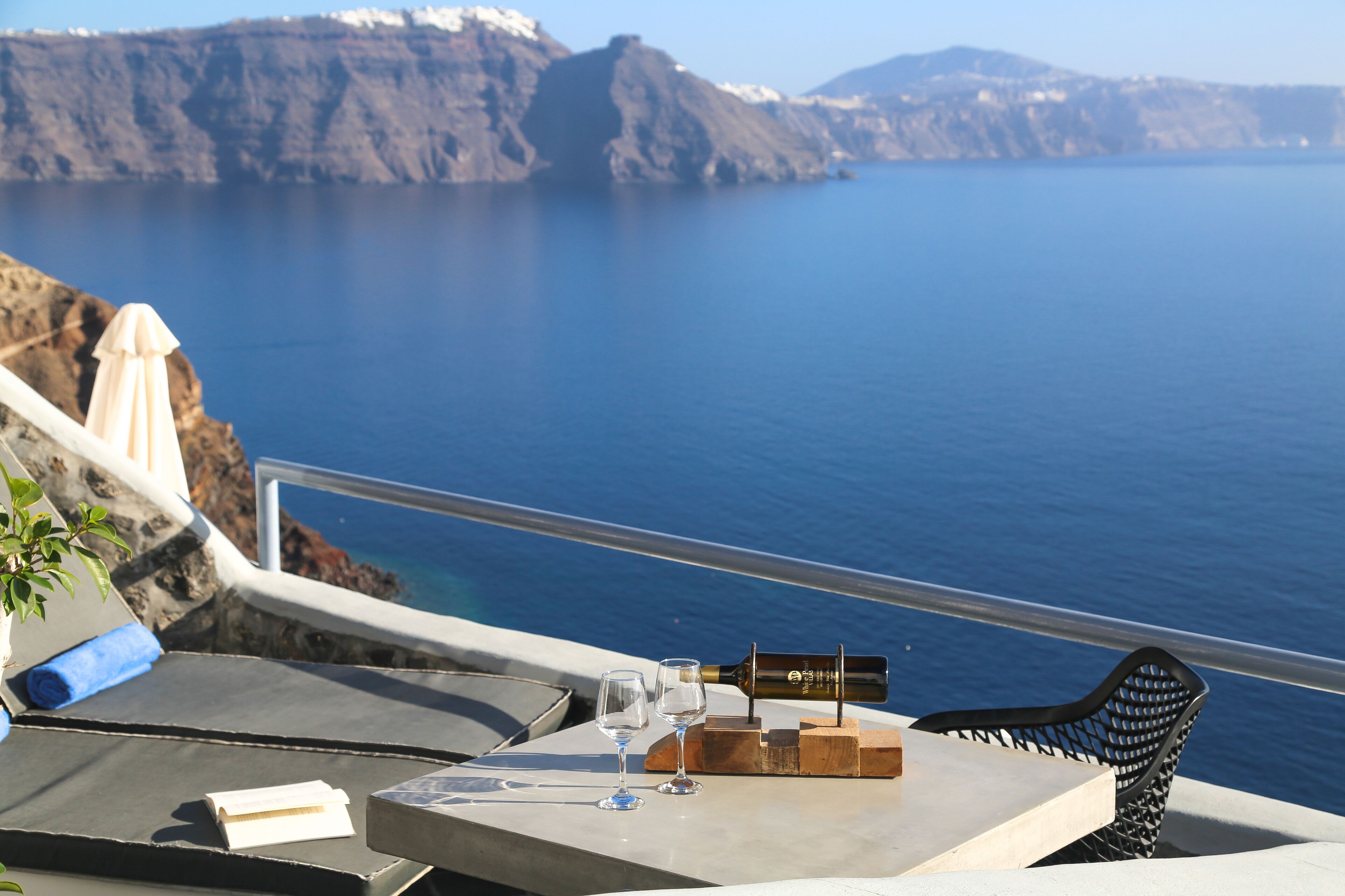A table with a bottle, two glasses, and a wooden block on a balcony overlooking a sea with cliffs.