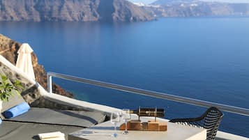 A table with a bottle, two glasses, and a wooden block on a balcony overlooking a sea with cliffs.