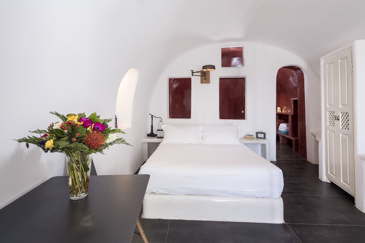 Un dormitori amb un llit, una tauleta de nit, un vas de flors i un passadís que condueix a una altra habitació.