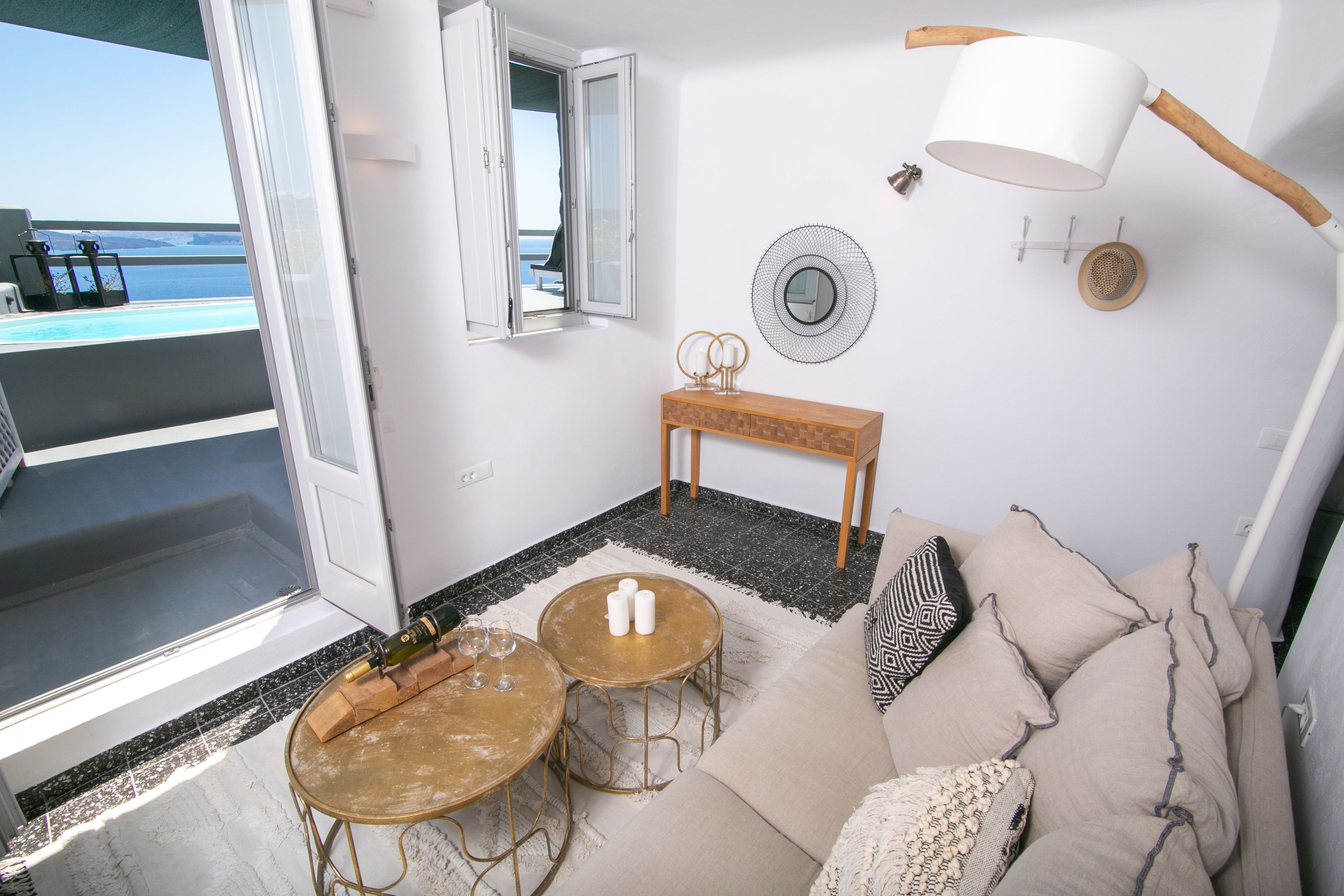 Une pièce comprenant un canapé, deux tables rondes, un banc en bois, une lampe fixée au mur, un miroir et une vue sur la mer par une porte ouverte.