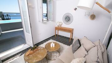 Una habitación con sofá, dos mesas redondas, un banco de madera, una lámpara de pared, un espejo y vista al mar a través de un puerta abierta.