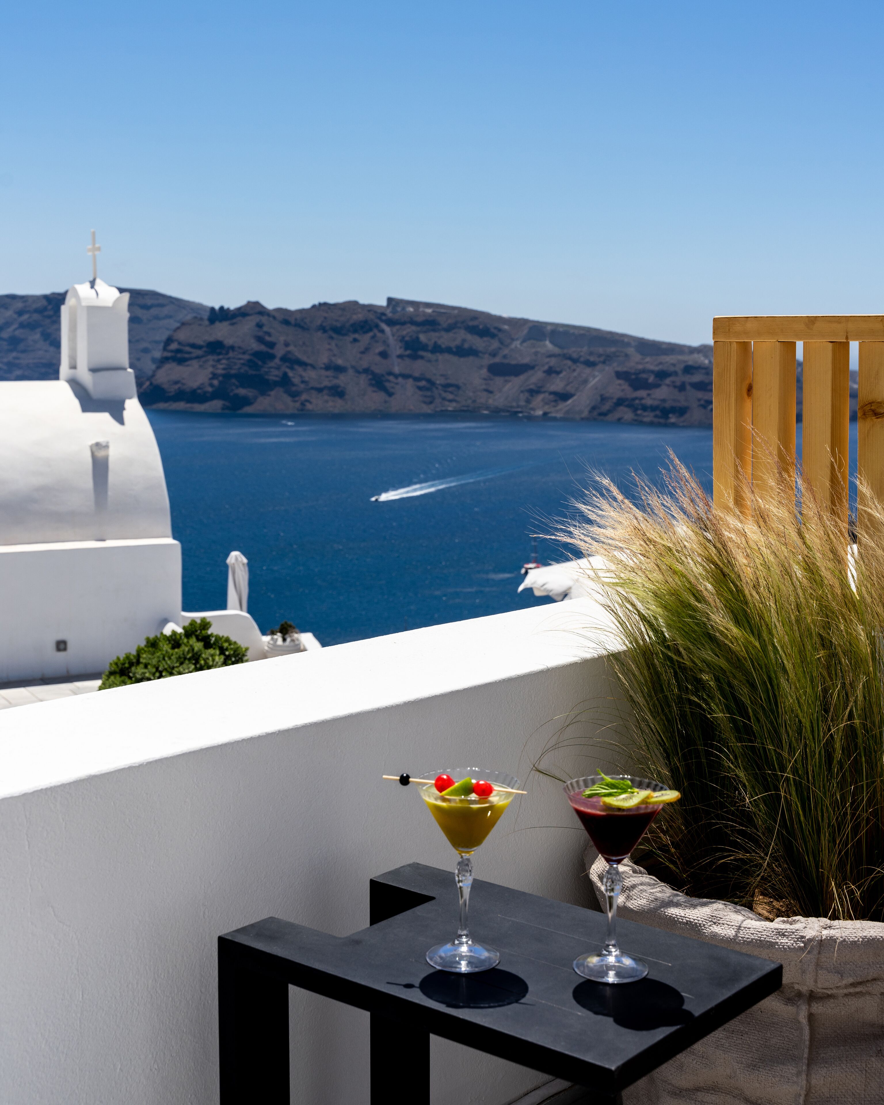 A table with two drinks and a view of the sea and white buildings.