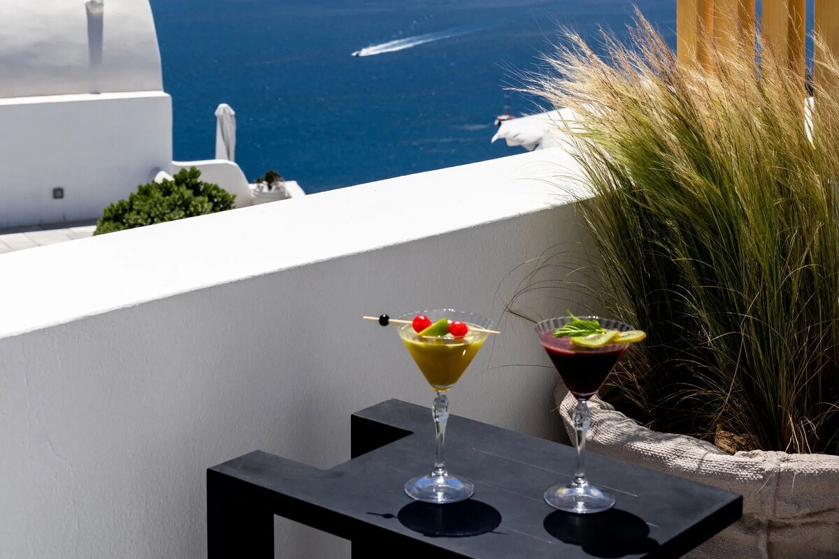 Un balcón con vista al mar y un edificio blanco con una cruz en la parte superior. Hay dos vasos de bebidas sobre una mesa.