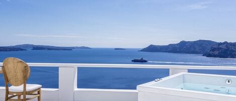 A balcony with a white bathtub, a wooden chair, and towels, overlooking a clear blue sea and distant islands.