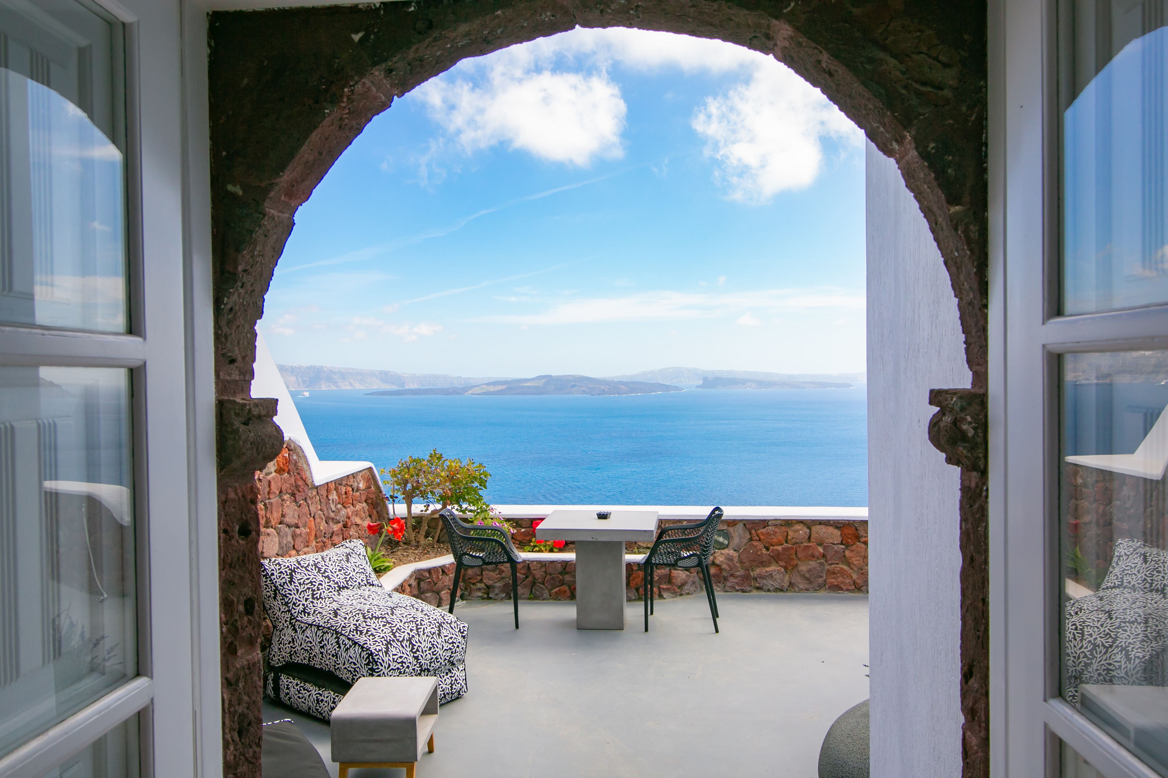 Vista a través de un arco hacia un patio con mesa y sillas, con vista a un paisaje costero.