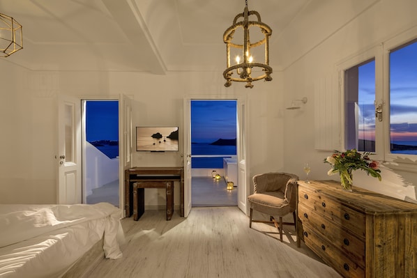 Un dormitorio con cama, cómoda de madera, silla y escritorio. Hay dos puertas que dan a un balcón con vista al cielo.