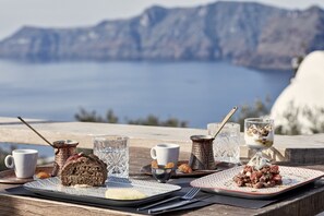 Mesa con comida que incluye pan, yogur y un postre, con vista panorámica a un cuerpo de agua y montañas.