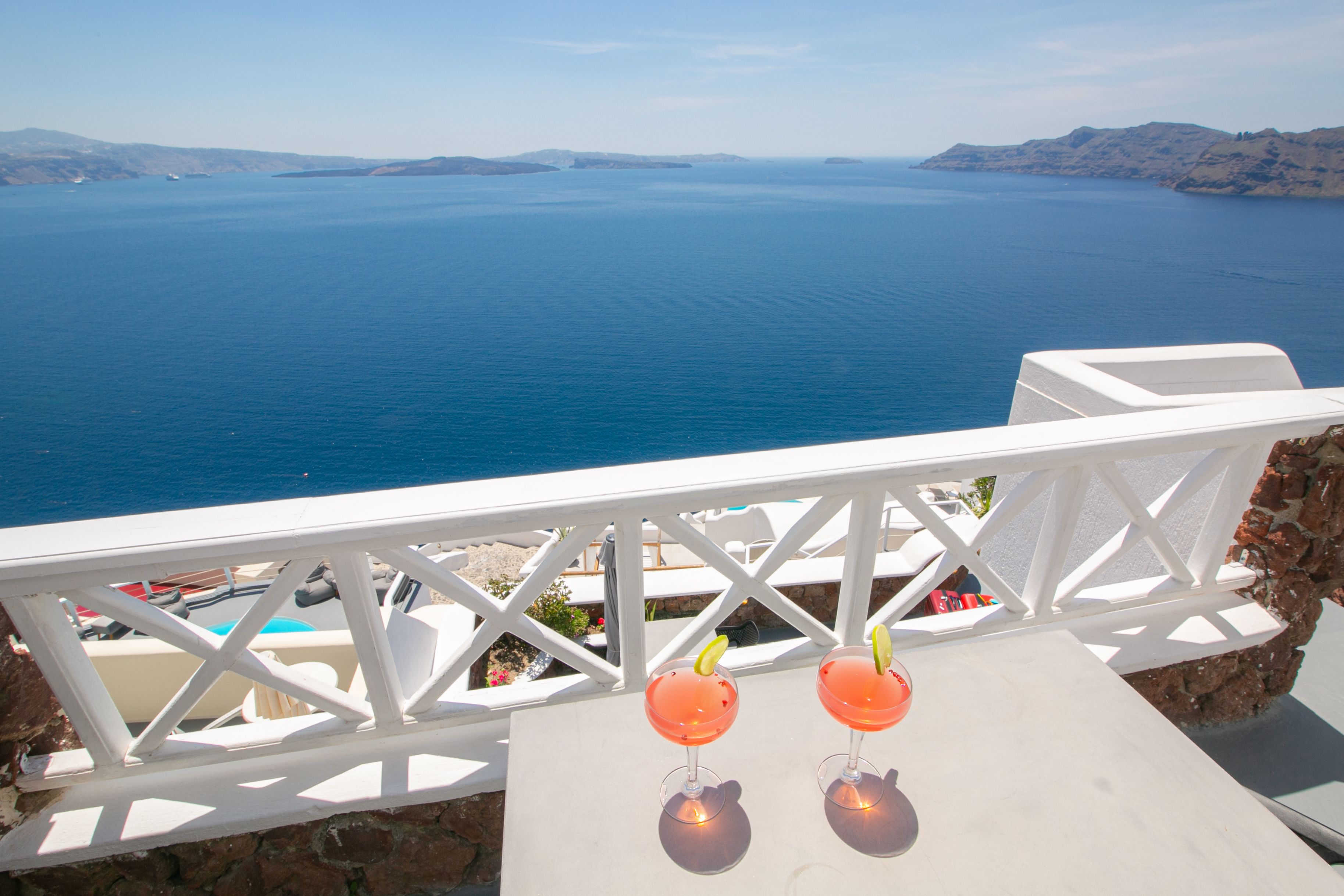 A balcony with a view of the sea and distant islands. There are two glasses of red drink on the table.