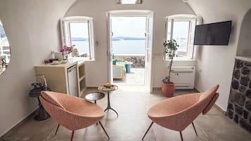 Un espacio habitable pequeño con dos sillones rosa, una mesita, un televisor montado en la pared y vista al mar a través de puertas abiertas.
