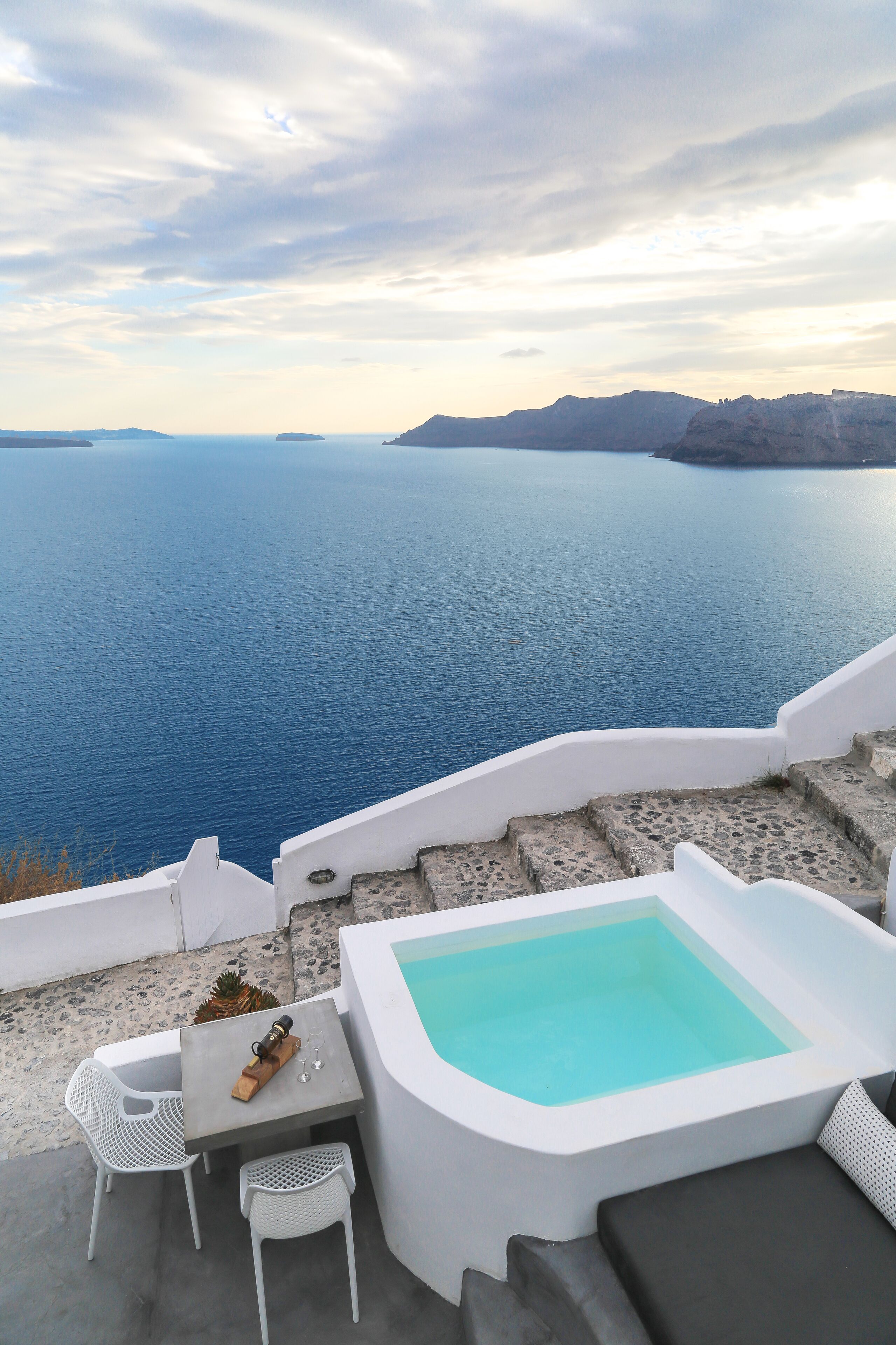 A pool area with a view of the sea, white outdoor furniture, and a table with a bottle and glasses.
