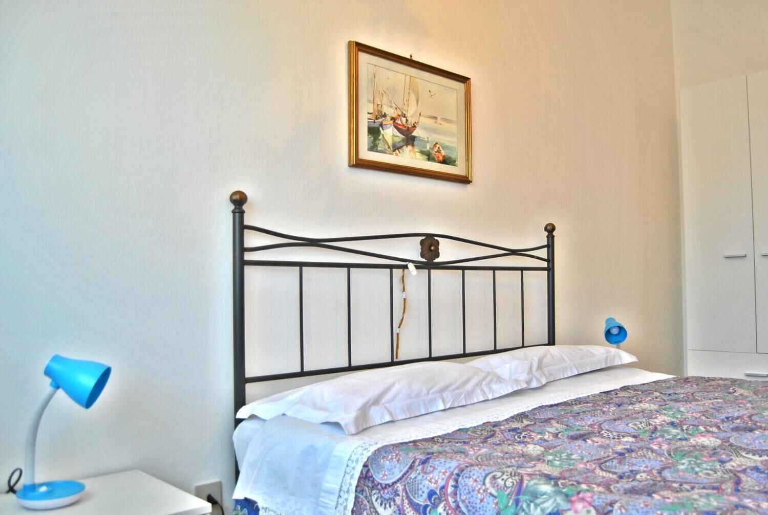 A bedroom with a metal bed frame, white bedding, a patterned rug, a blue bedside lamp, and a framed picture on the wall.
