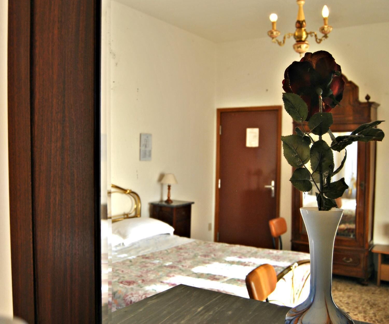 A bedroom with a bed, a nightstand, a lamp, a dresser, and a vase with a rose.
