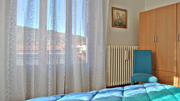 A bedroom with a bed, a radiator, a window with curtains, a wardrobe, and a painting on the wall.