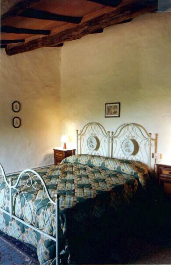 Un dormitorio con estructura de cama metálica, colcha estampada y una sola mesita de noche con lámpara.
