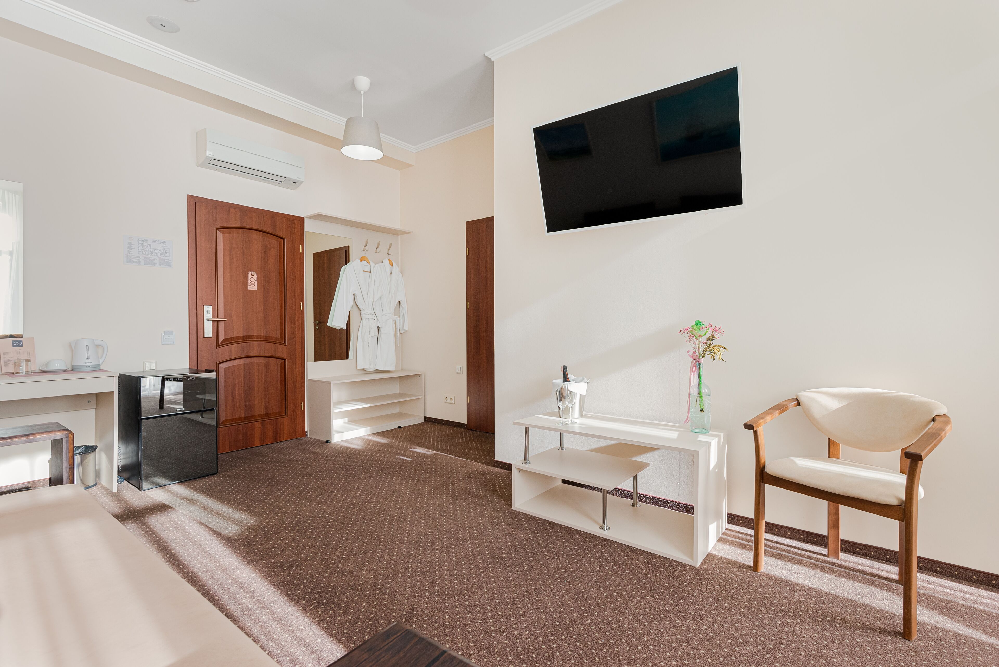 Habitación de hotel con kitchenette, televisor de pantalla plana montado en la pared, una silla de madera y una mesa pequeña con un jarrón de flores.