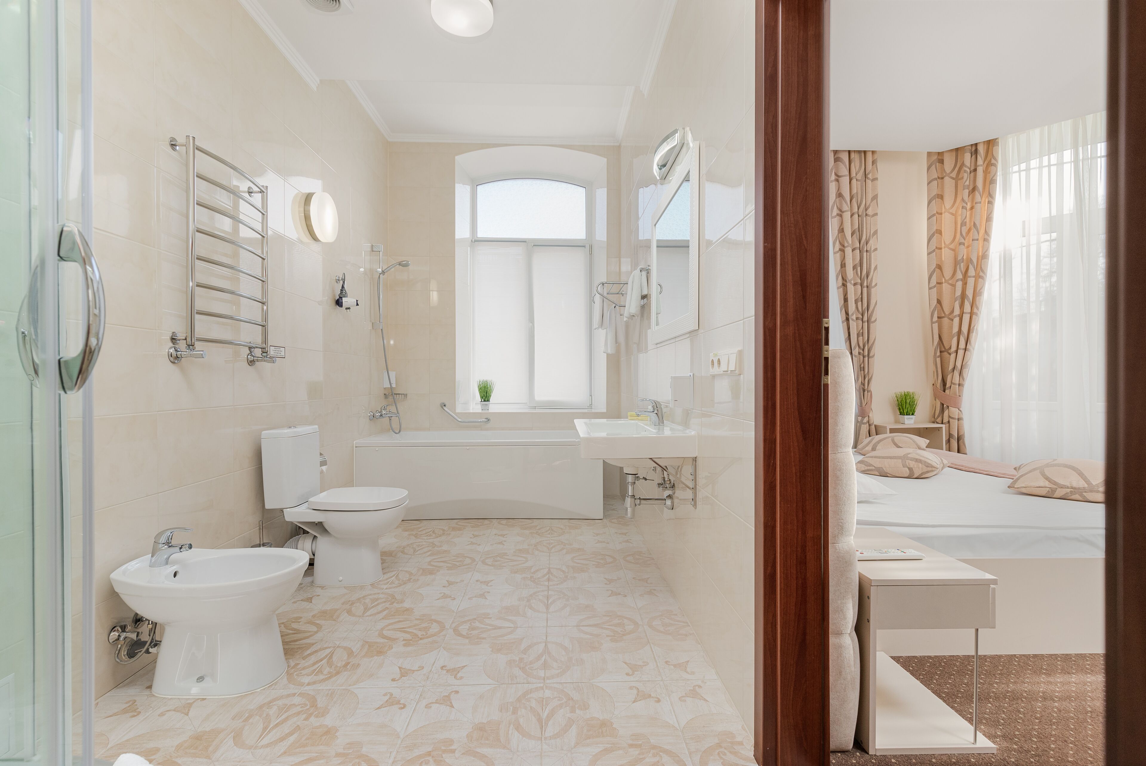 A modern bathroom with a bidet, toilet, and bathtub. It features a large mirror, a sink, and a window with curtains.