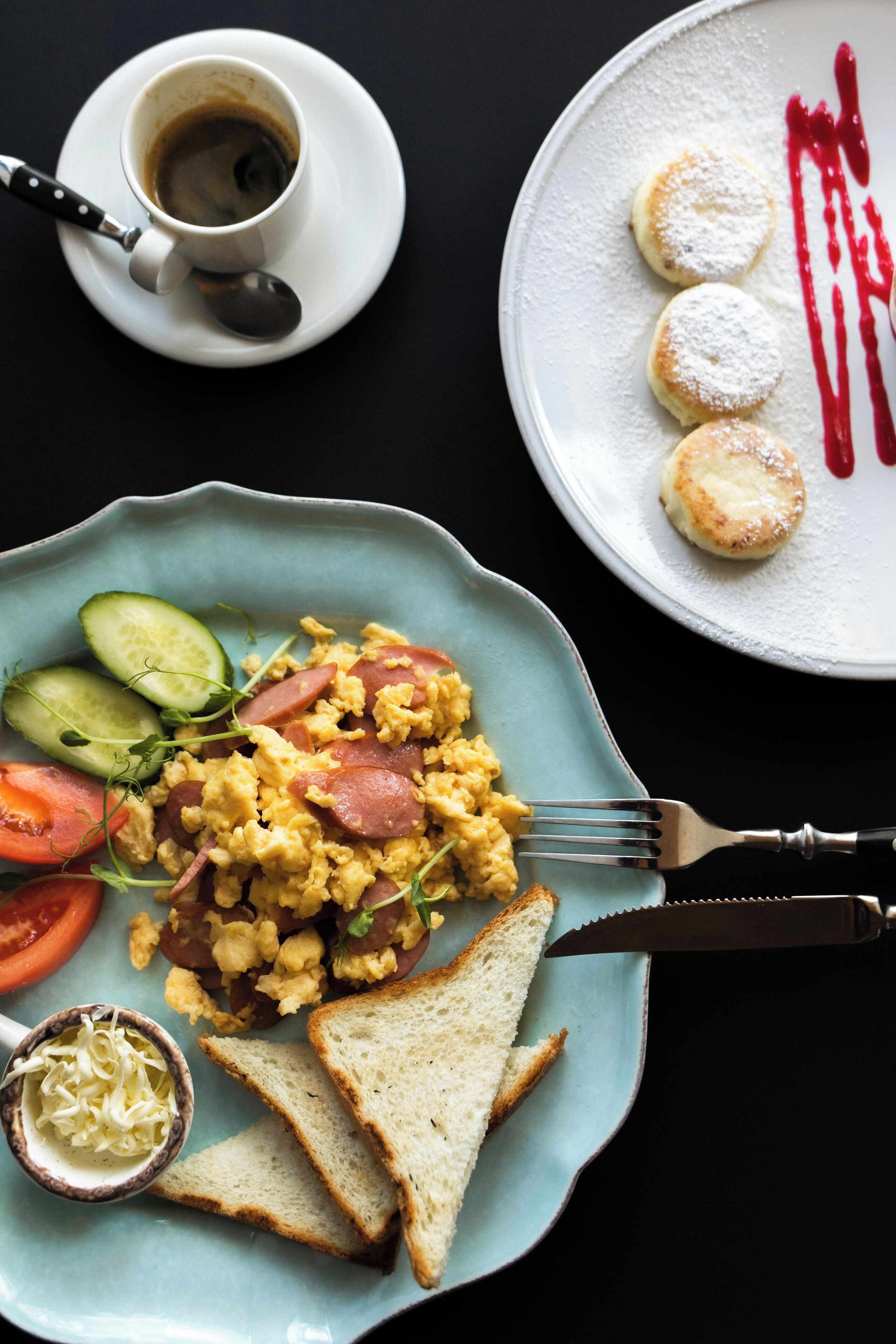 A breakfast plate with scrambled eggs, sausage, cucumber slices, and toast, a cup of coffee, and a plate with pastries and powdered sugar.