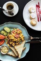 Ontbijtplaat met roerei, worst, augurkplakjes en toast, een kop koffie, en een bord met gebak en poedersuiker.