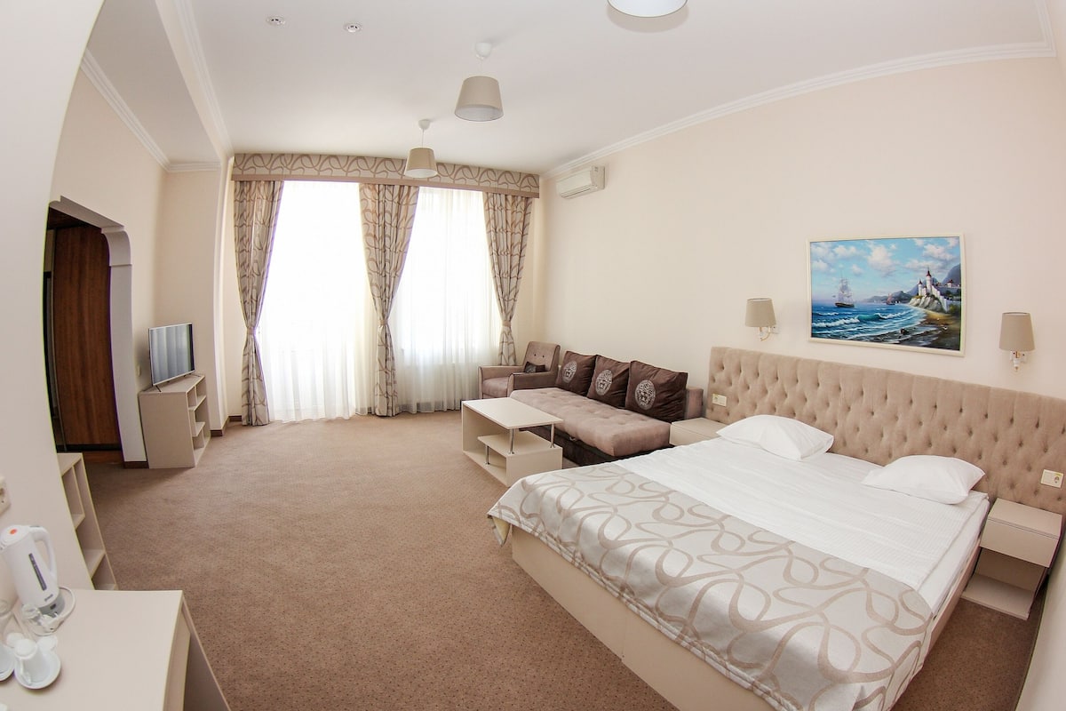 A hotel room with a large bed, a sofa, a desk with a lamp, and a painting on the wall.