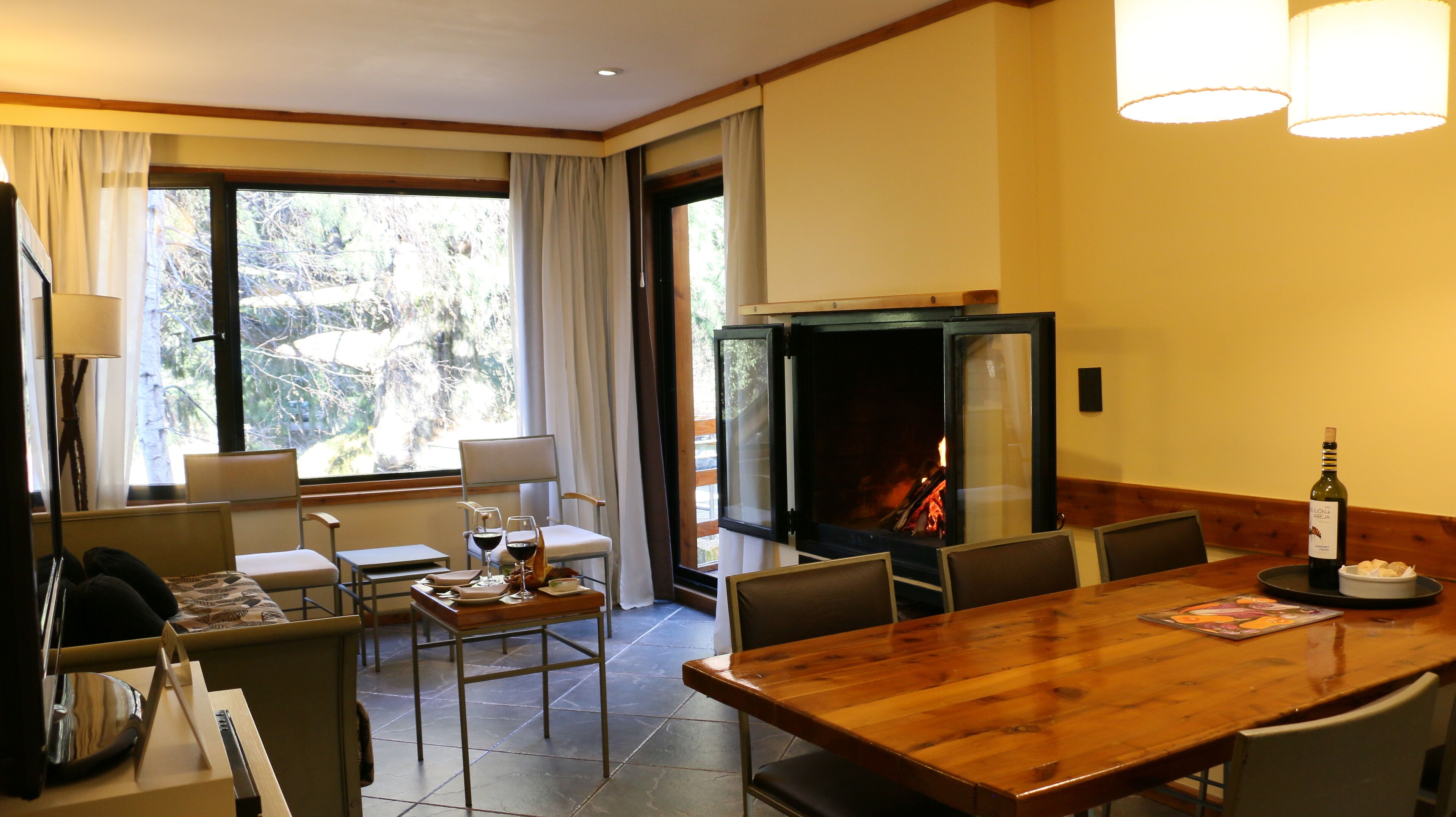 A dining area with a wooden table and chairs, a fireplace, and a view of the outdoors through large windows.