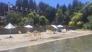 A beach with sun loungers and umbrellas, a wooden building in the background, and a clear view of the water. - Bahia Manzano Resort (Villa La Angostura)