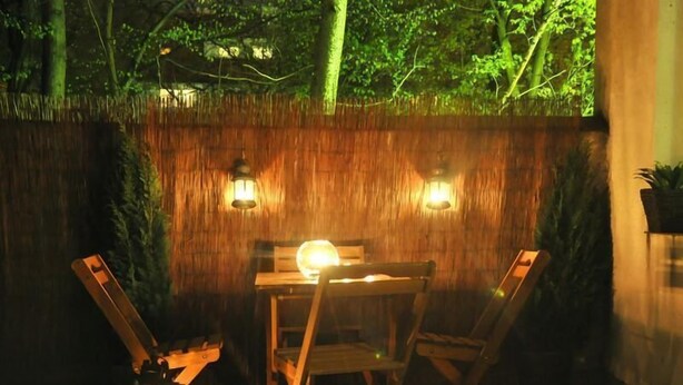 An outdoor dining area with a wooden table and chairs, two wall-mounted lanterns, and a thatched wall backdrop. - Anton House (Warsaw)