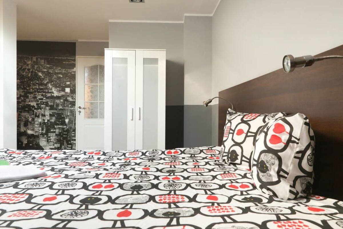 A bedroom with a patterned bedspread, a cityscape wall, a white wardrobe, and a dark headboard.