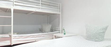 A bunk bed room with a white color scheme, a small bedside table with a green vase, and a patterned pillow.