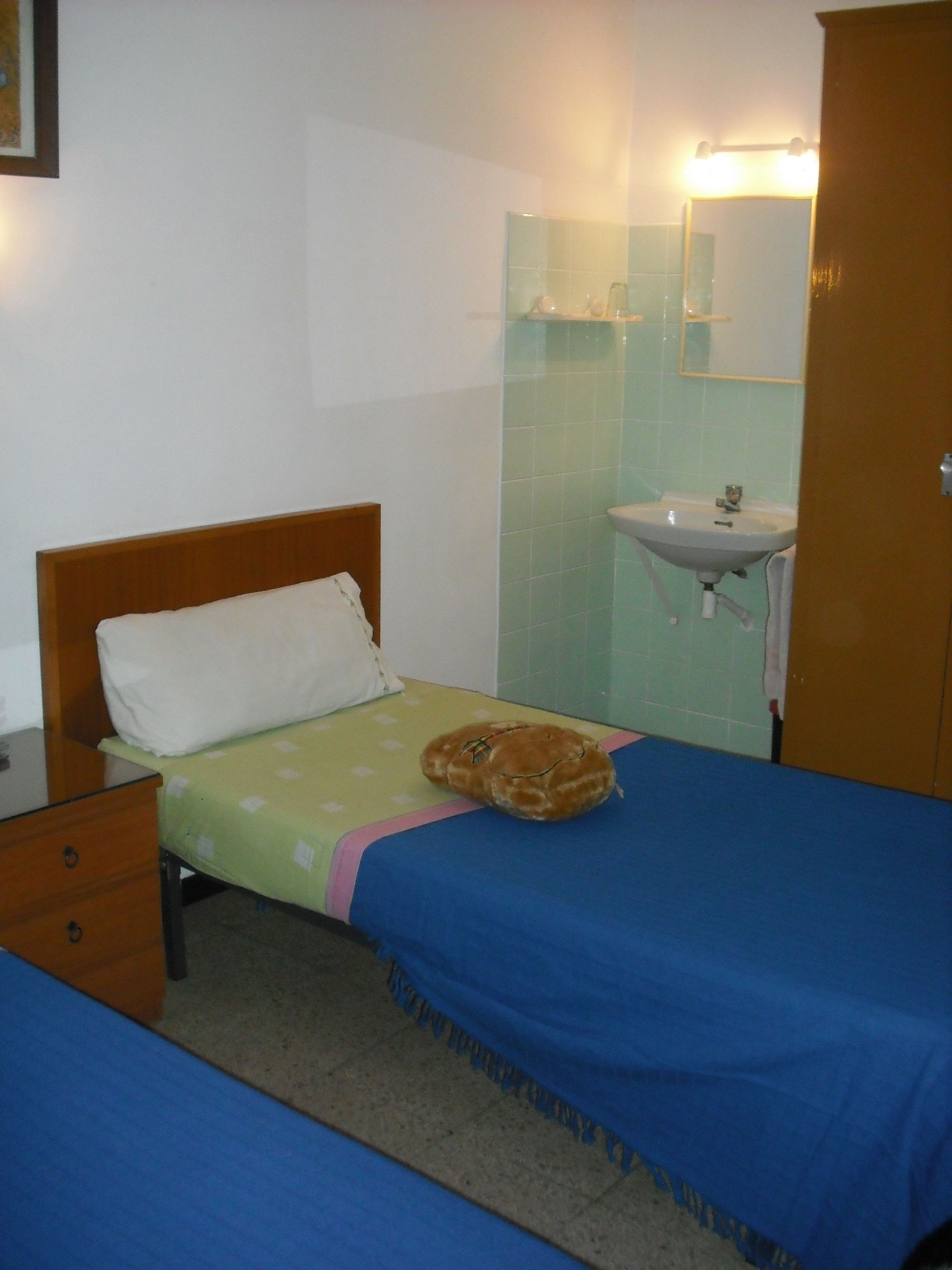 Una habitación pequeña y sencilla con cama, mesita de noche, un ventanal y baño con lavamanos y espejo.