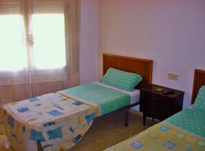 A room with two single beds, a small bedside table with a lamp, a window with curtains, and a wooden floor.
