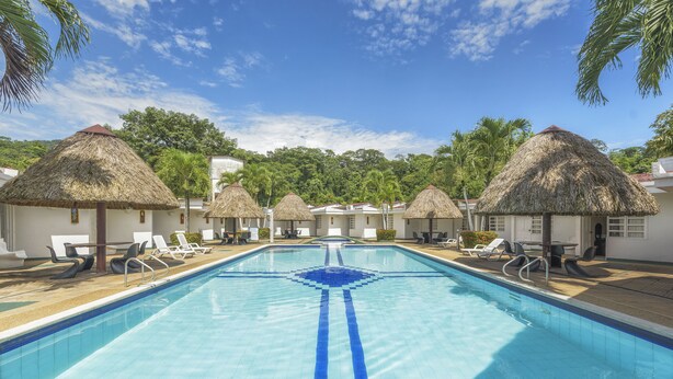 A swimming pool with lounge chairs and thatched-roof huts, surrounded by palm trees and greenery. - Hotel Santa Barbara (Villavicencio)