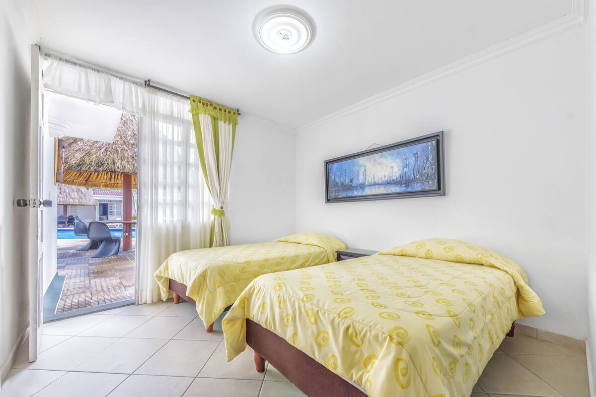 A room with two beds, a painting on the wall, and a view of a pool area through the door.