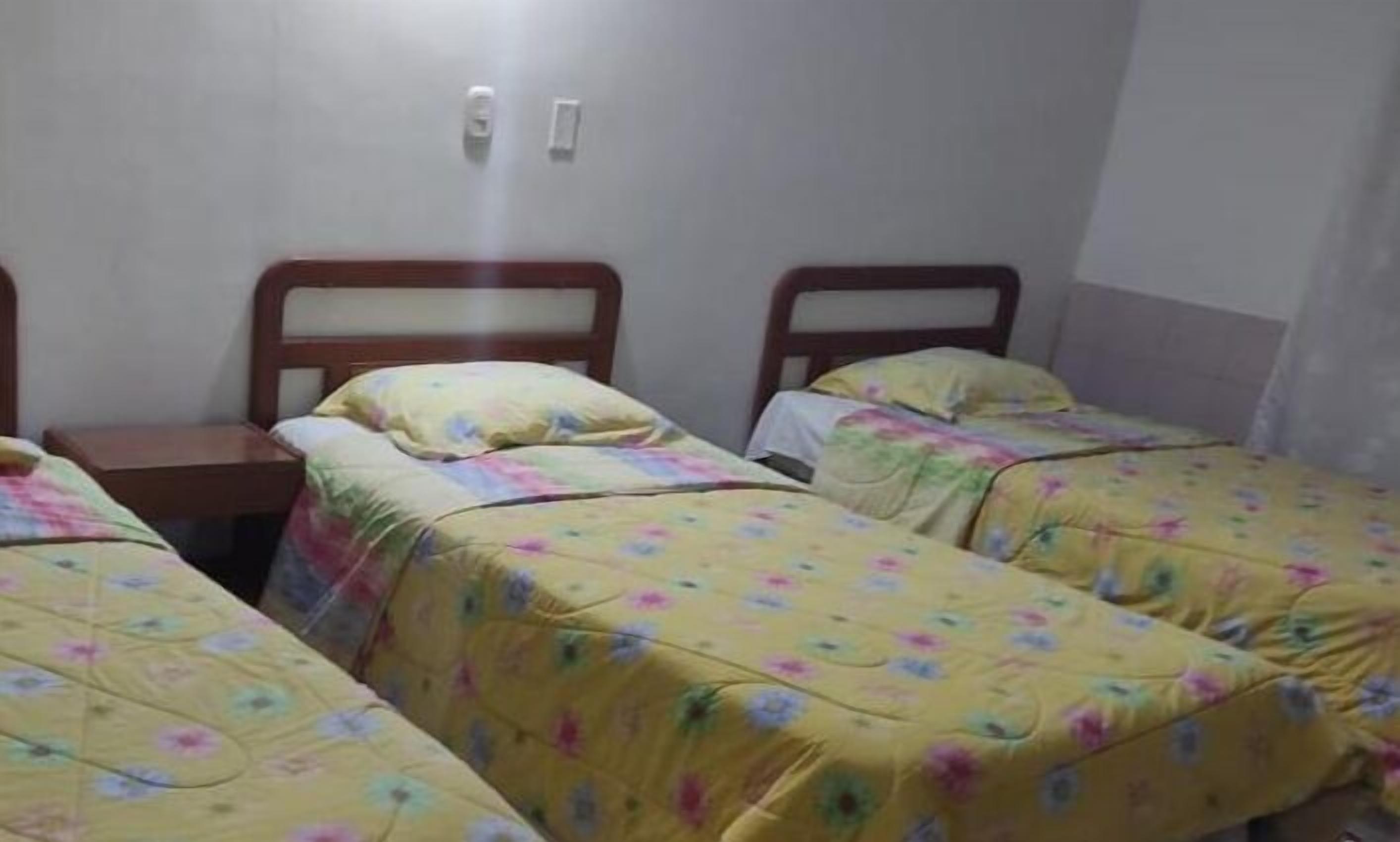 A room with two single beds, each with a wooden headboard and floral bedding. There is a small wooden nightstand between the beds.