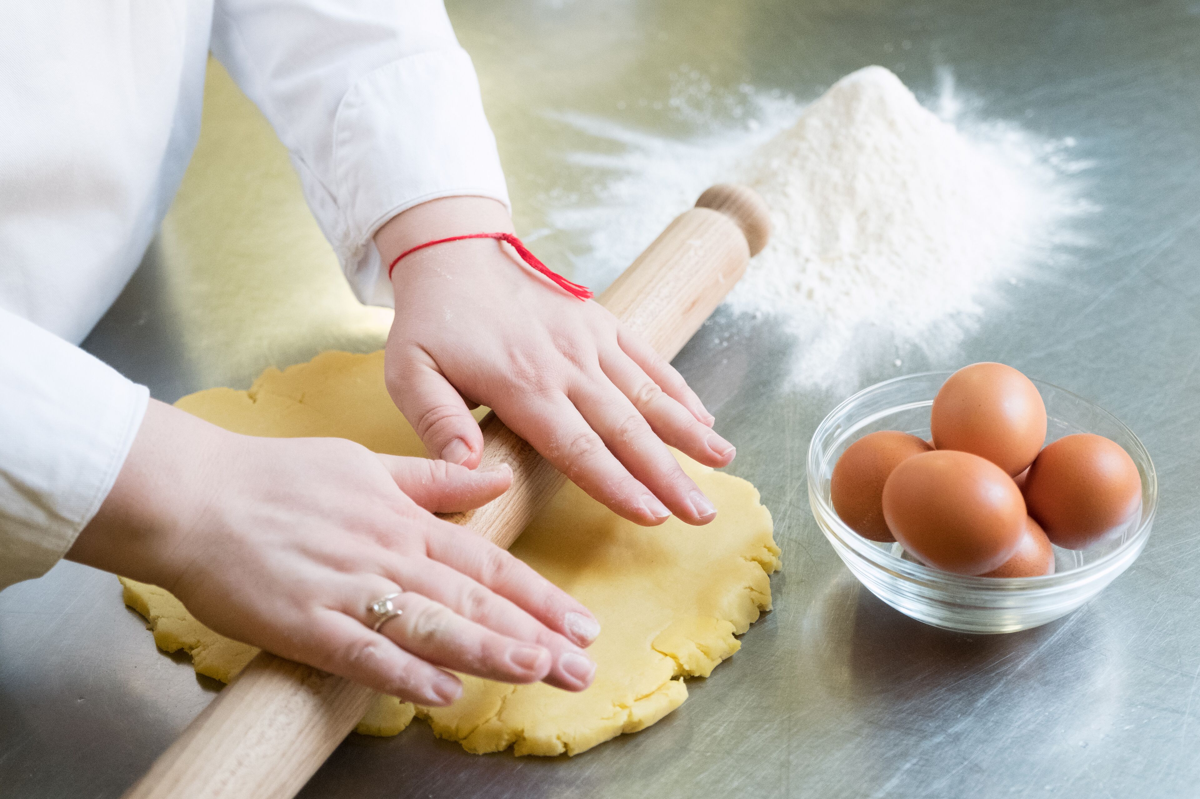 A person rolling dough with a rolling pin on a metallic surface, a bowl of eggs, and a pile of flour nearby.
