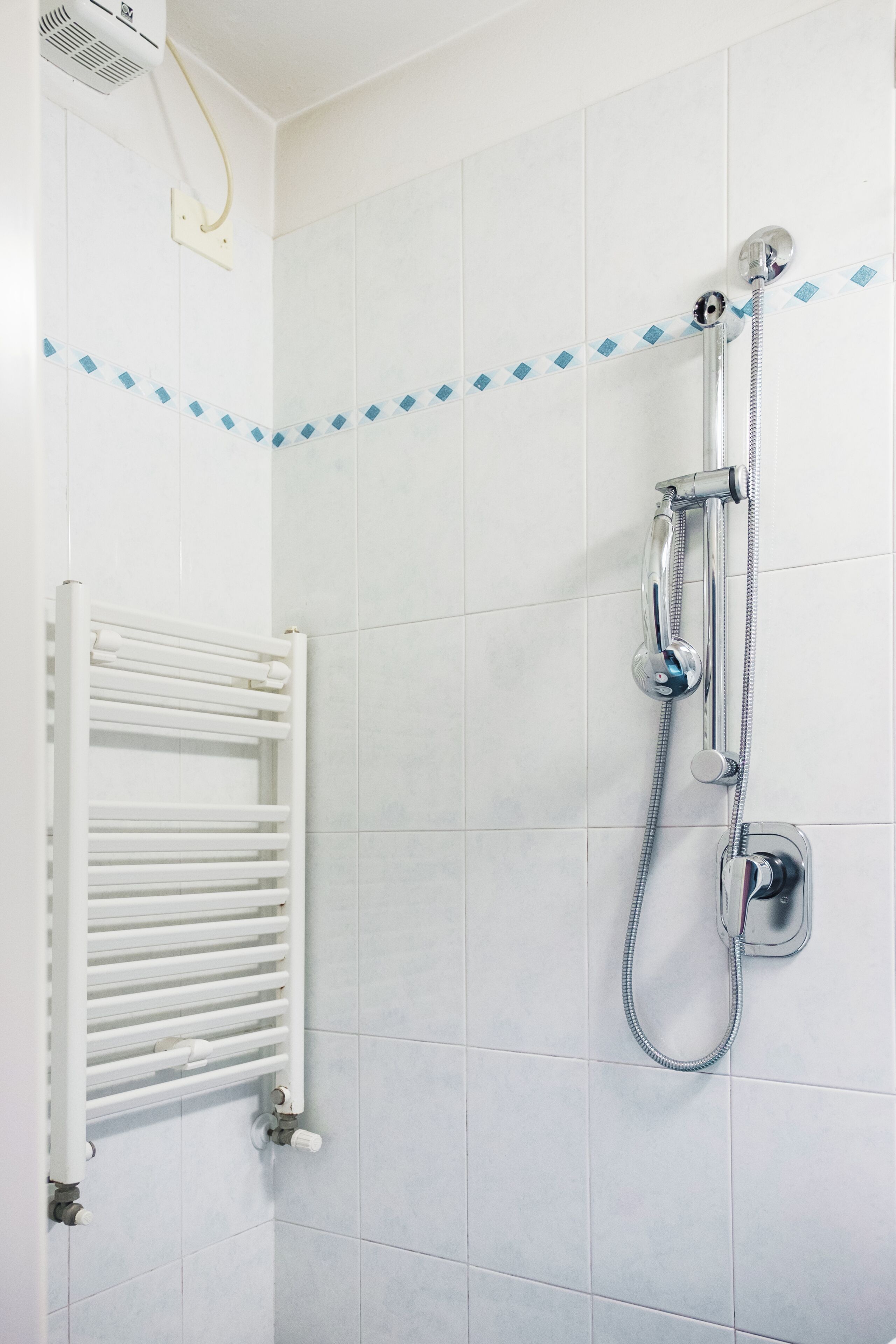 A shower with a handheld hose and a wall-mounted showerhead, a white towel rack, and a tiled shower area.