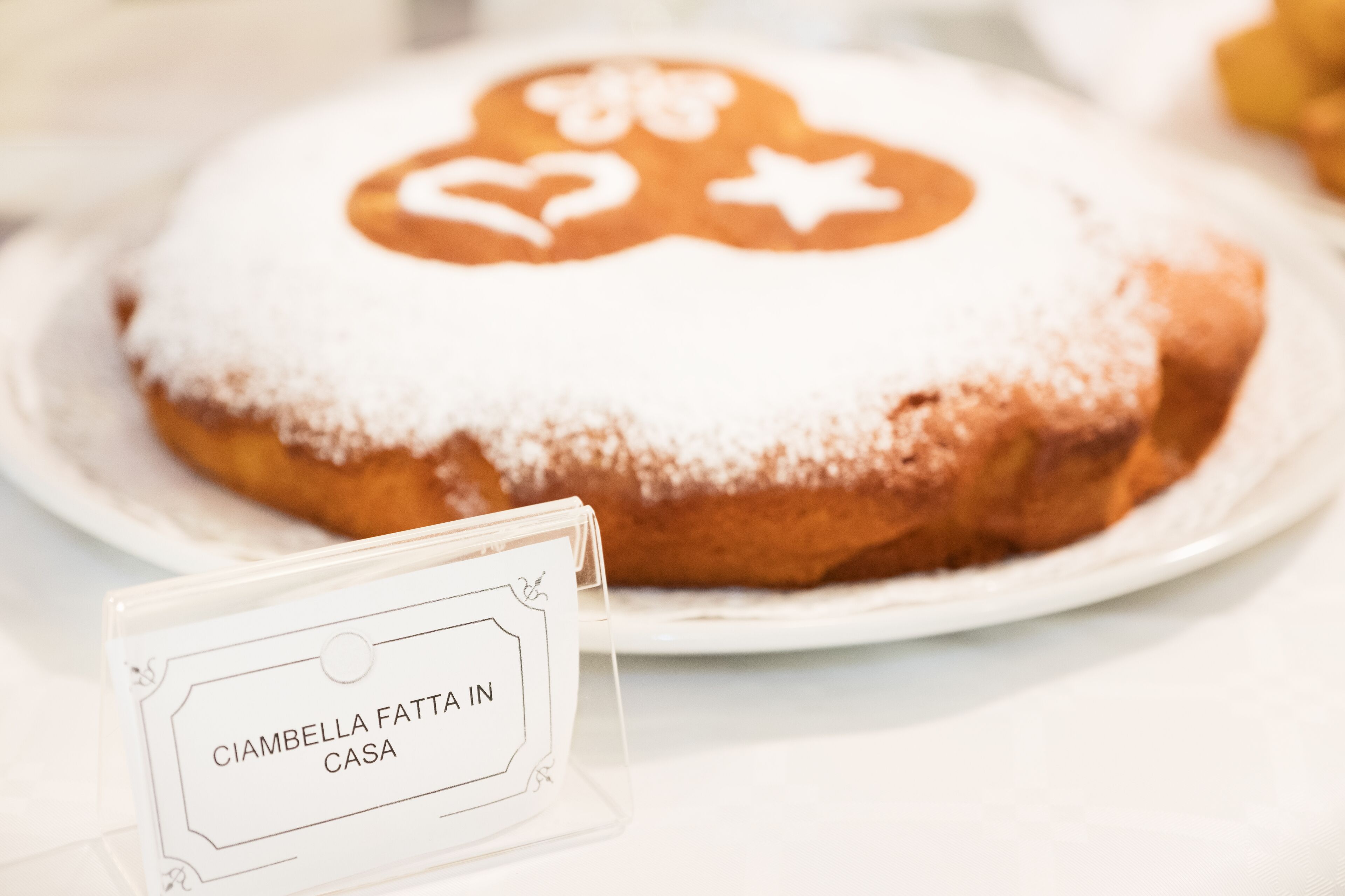 Valkoisella lautasella on kakku, jossa on etiketti, jossa lukee: ”CIAMBELLA FATTA IN CASA”.