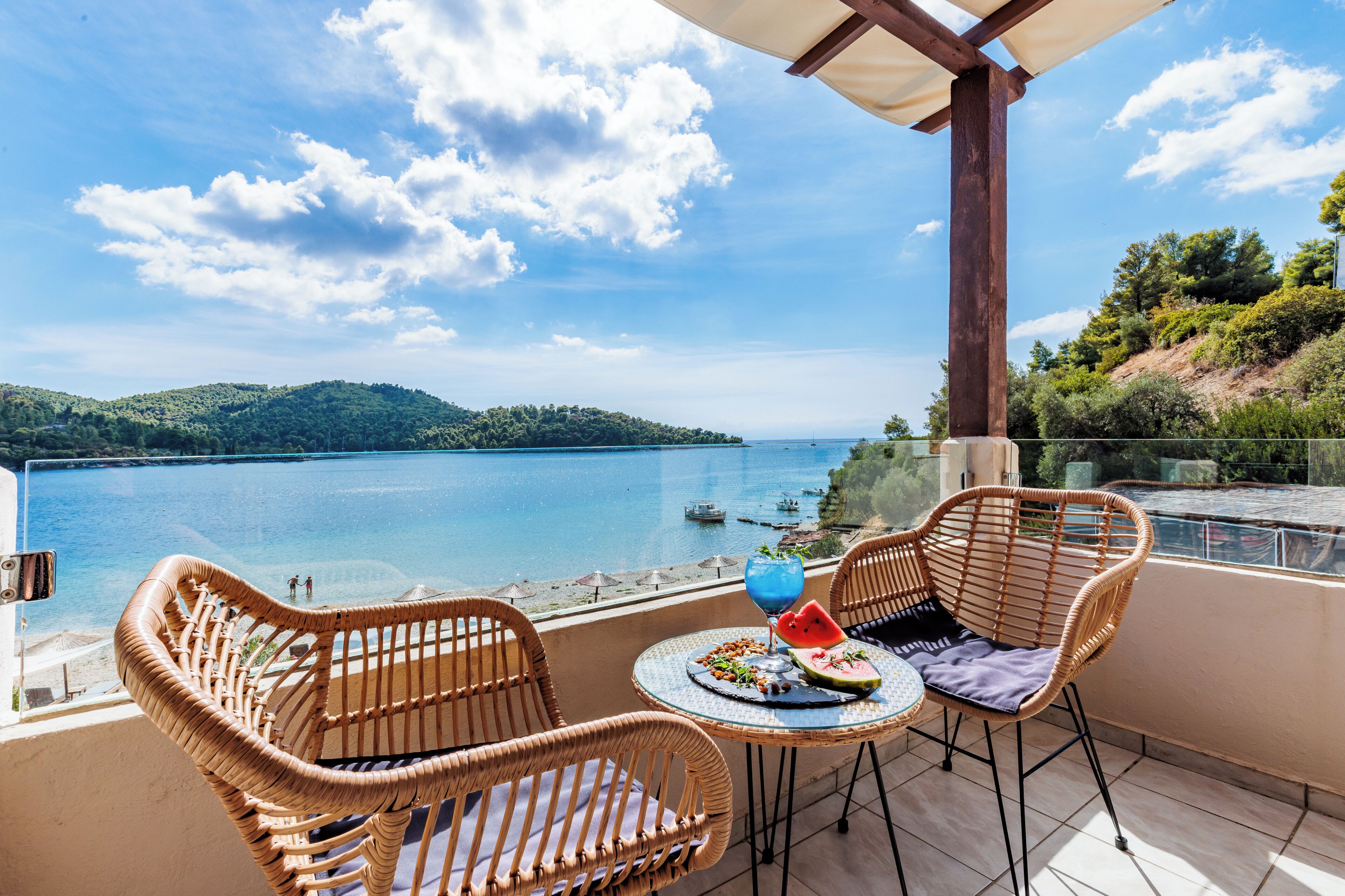 Un balcon avec des fauteuils en osier, une table en verre et une vue sur un lac et des collines.
