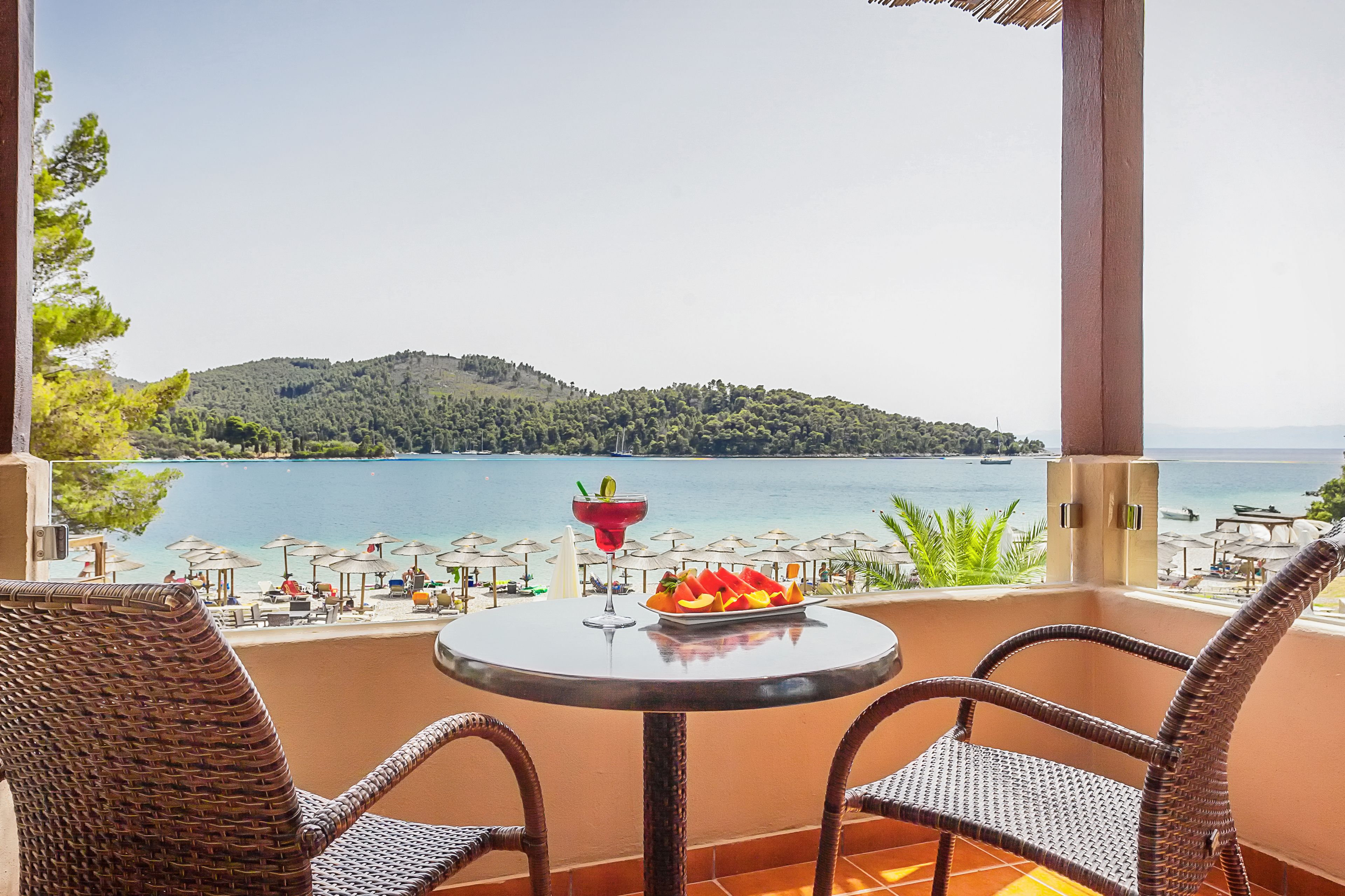 Un balcon avec des fauteuils en osier, une table sur laquelle est disposée une assiette de fruits et une boisson, et une vue sur une plage et la mer.