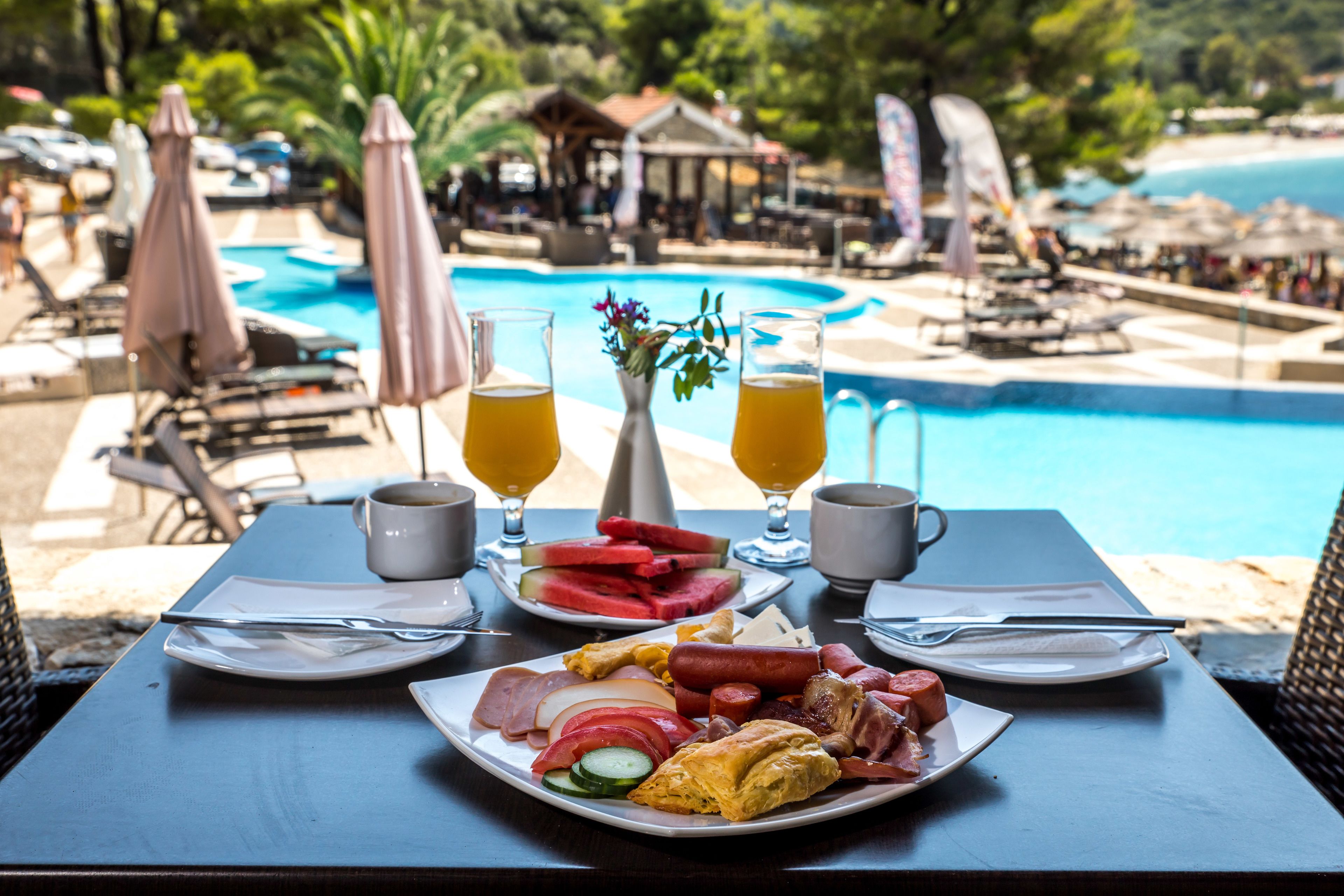 Une table à manger avec des aliments pour le petit-déjeuner, deux verres de jus d’orange, et en arrière-plan, une vue sur une piscine et une plage.