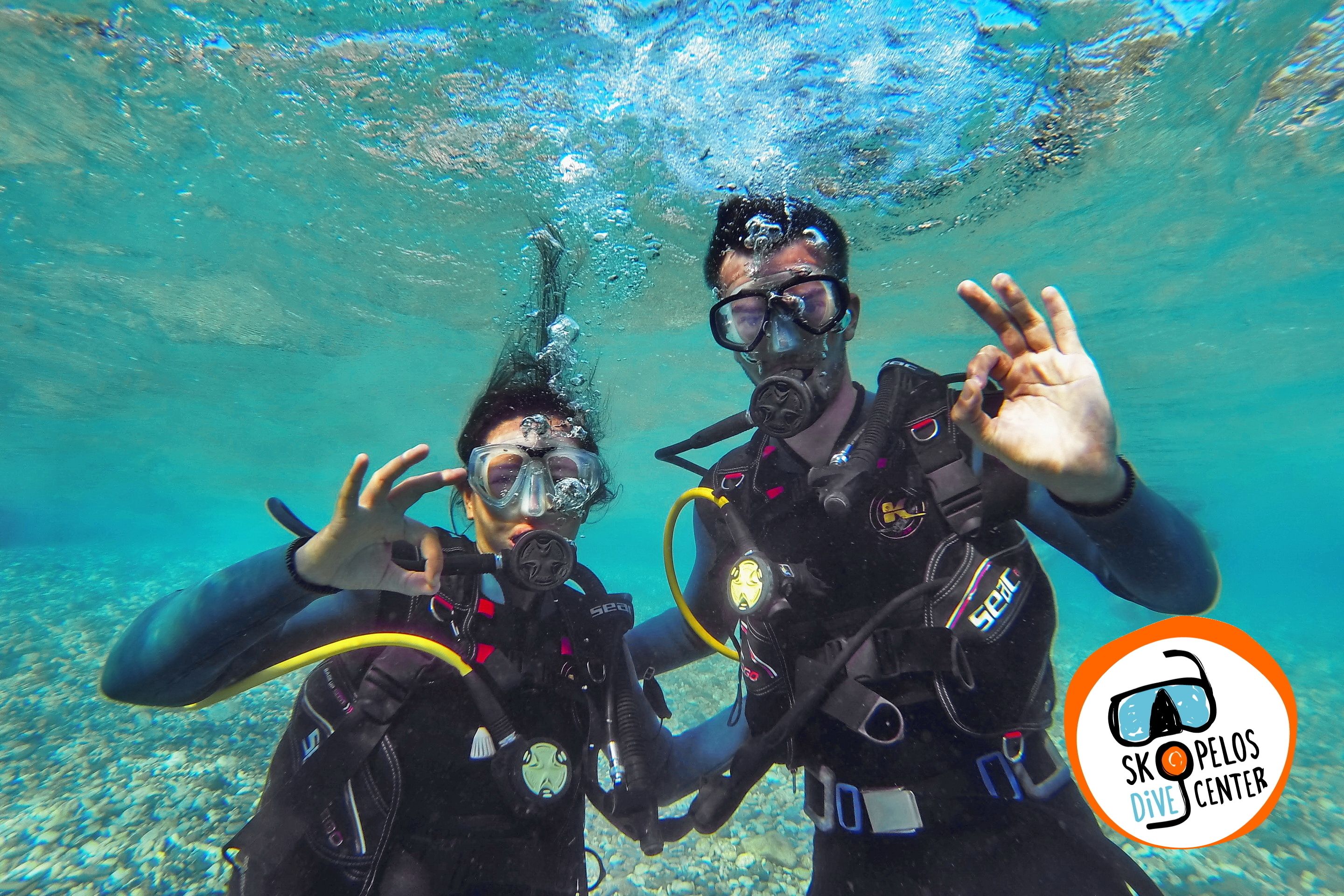 Two scuba divers underwater with a logo of 'SKOPELOS DIVE CENTER' in the bottom right corner.