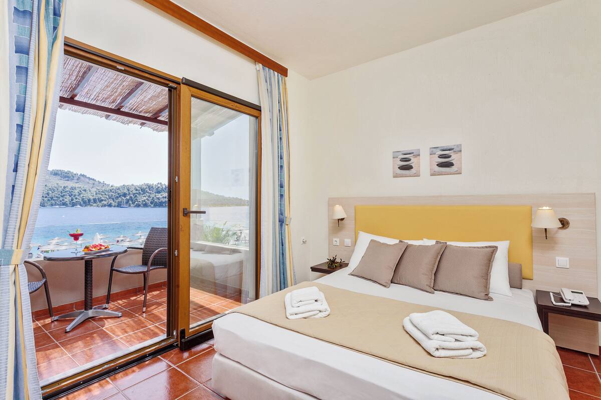 A hotel room with a bed, a small table with chairs, a view of the sea, and a balcony with a table and chairs.