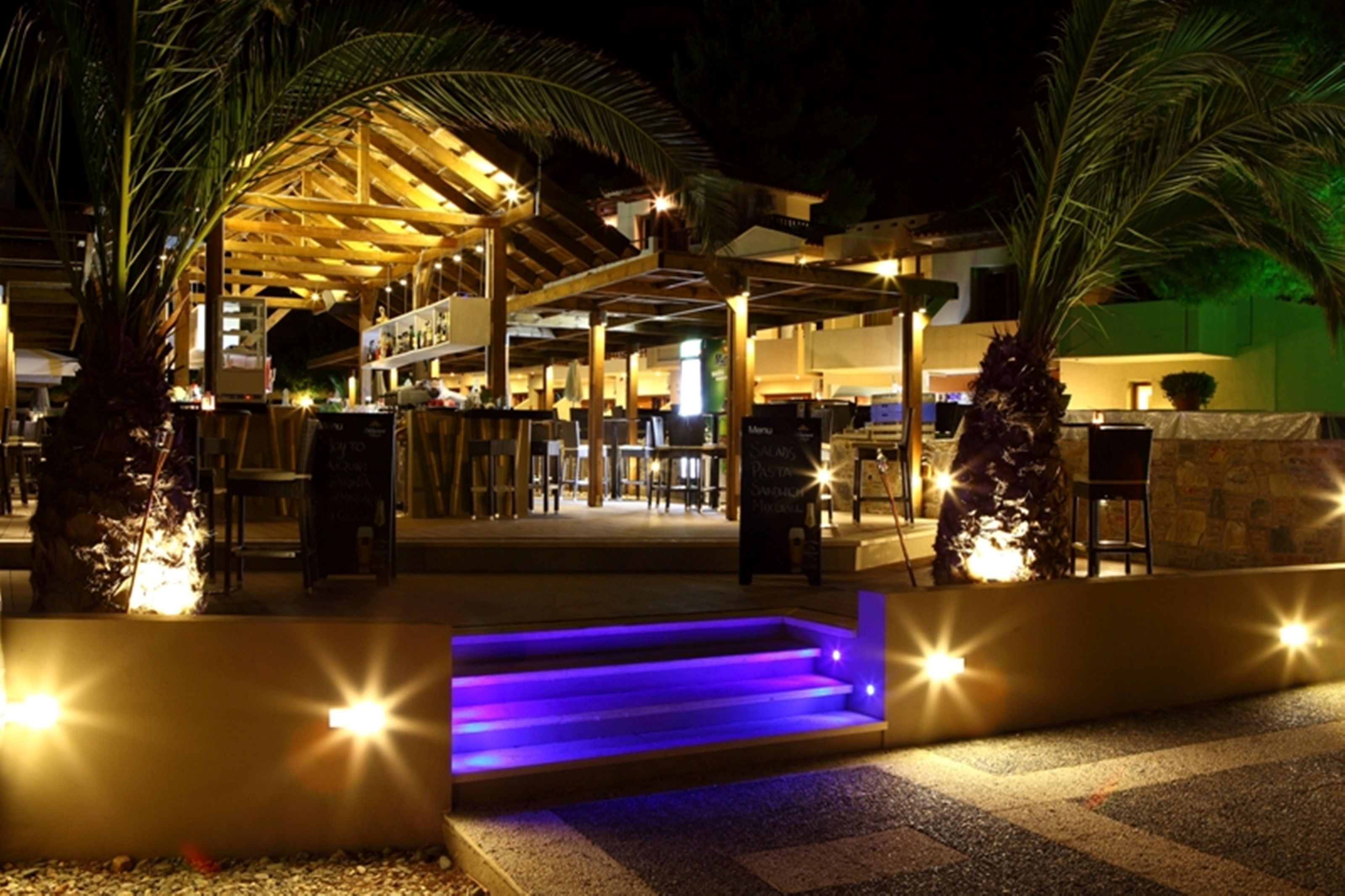 An outdoor area with a covered seating area, palm trees, and a staircase with blue lighting.