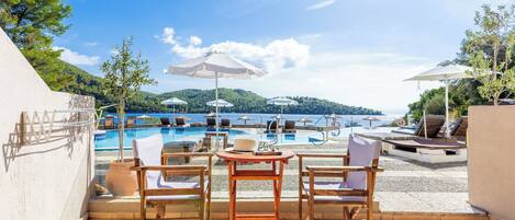 Zona de piscina con sillones y sombrillas, con vista a un mar azul y un paisaje montañoso.