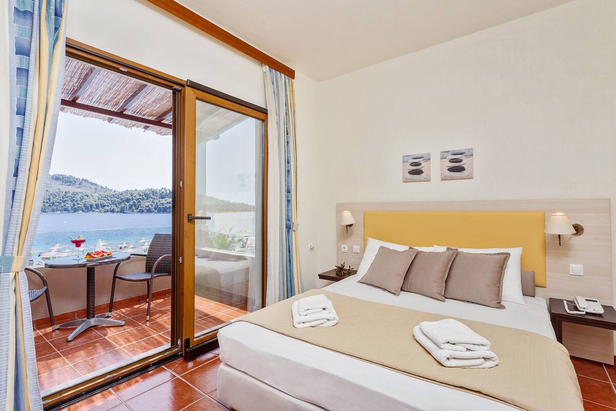 A hotel room with a bed, a small table with chairs, a view of the sea, and a balcony with a table and chairs.