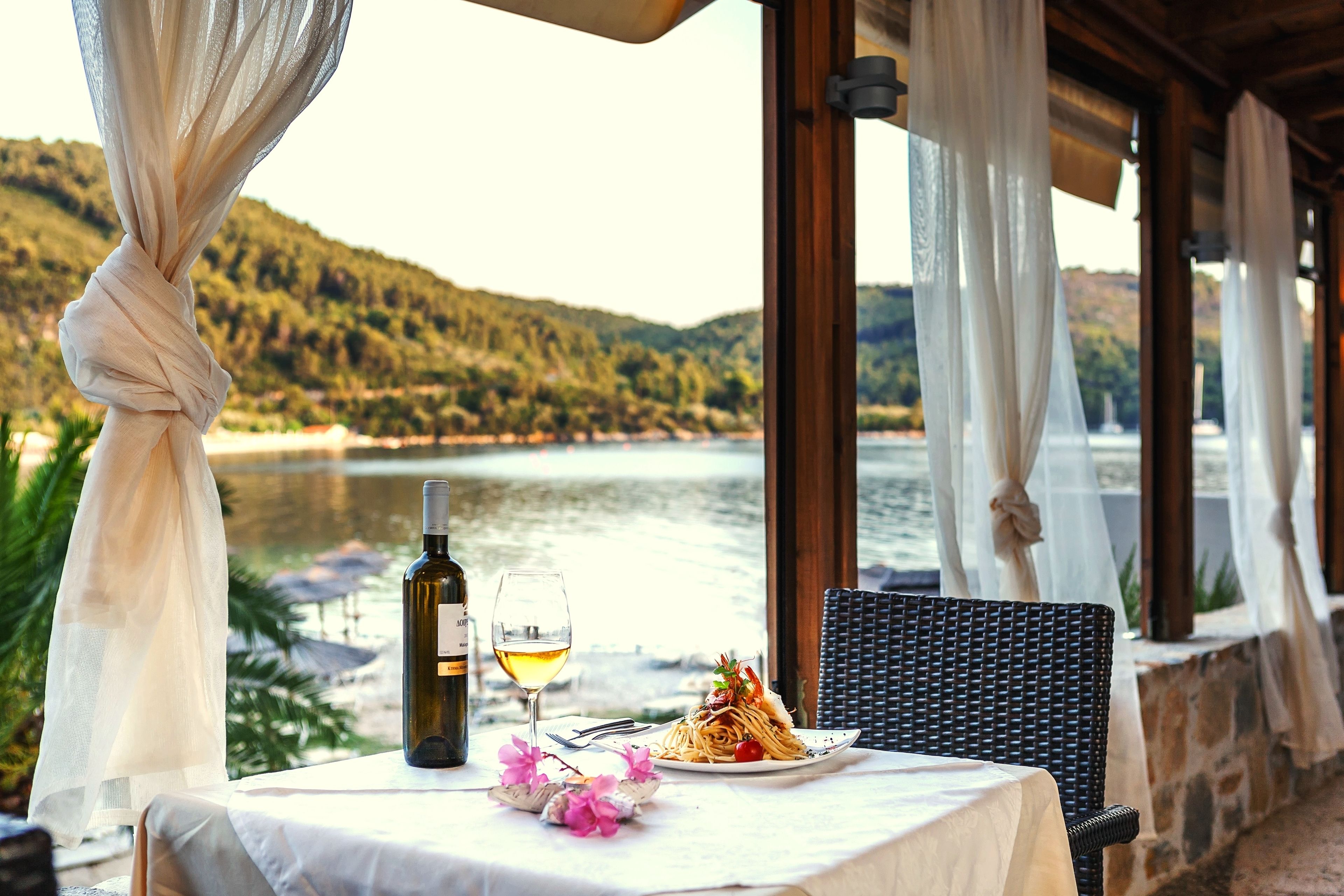 Una mesa con una botella de vino, una copa y un plato de comida, frente a una vista panorámica de un lago.