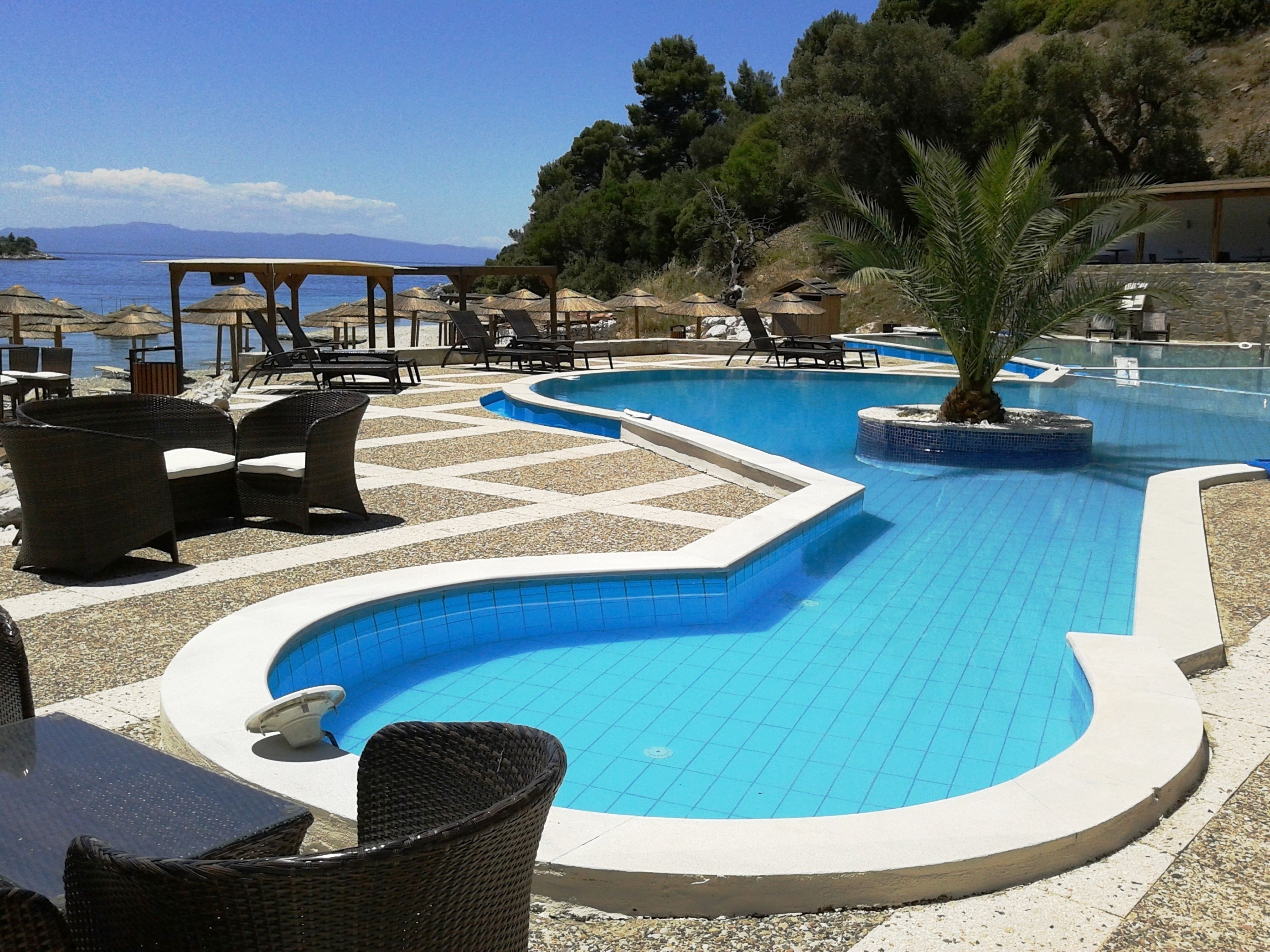 A pool area with lounge chairs and umbrellas, surrounded by a pebble beach and overlooking a body of water.